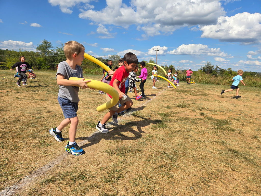 Several students run in different directions while others hold pool noodles, actively participating in a lively outdoor game.