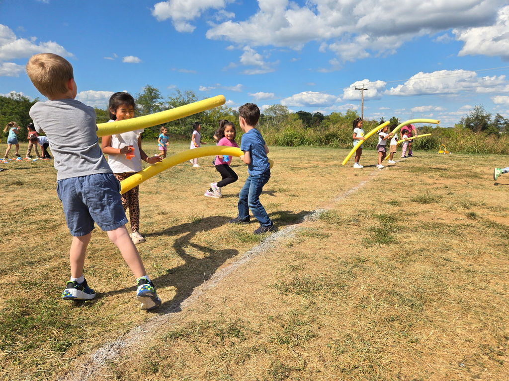 A group of children play an outdoor game on a grassy field, holding yellow pool noodles and using them to tag or block one another. Some students face off in the foreground while others run and play in the background under a bright blue sky with scattered clouds.