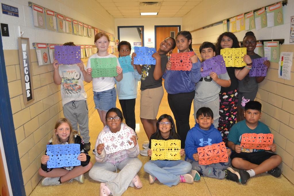 Image contains a class showing their papel picado artwork in the hallway.  Two rows of smiling students.