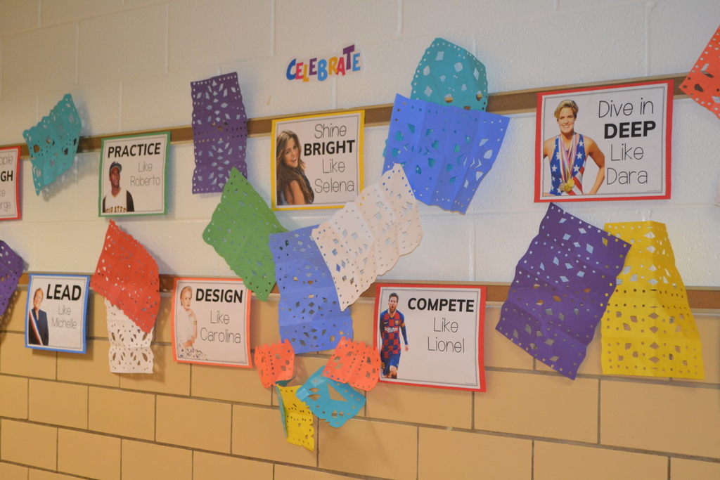 Image contains a school hallway display with papel picado artwork by students and signs that ready Practice like Roberto Shine Bright like Selena Compete like Lionel Dive Deep like Dara 