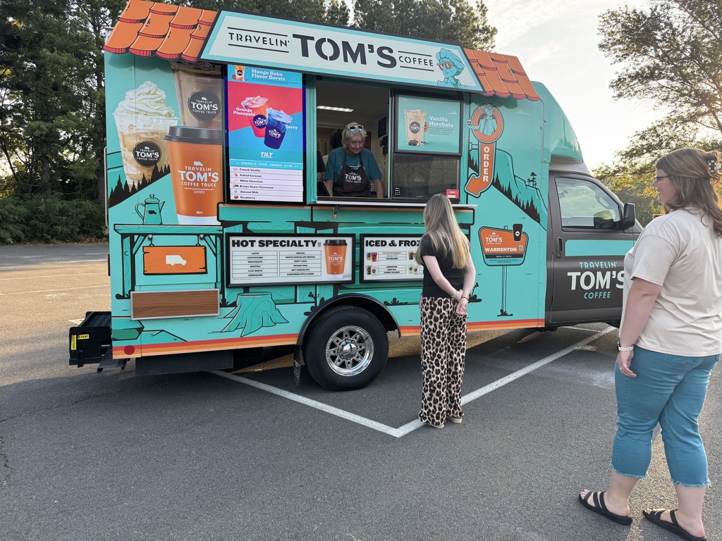 A brightly painted teal Travelin’ Tom’s Coffee Truck is parked in a lot. Two women stand at the window ordering drinks, while a worker inside the truck takes their order. The truck’s menu and large coffee graphics are visible on the side.