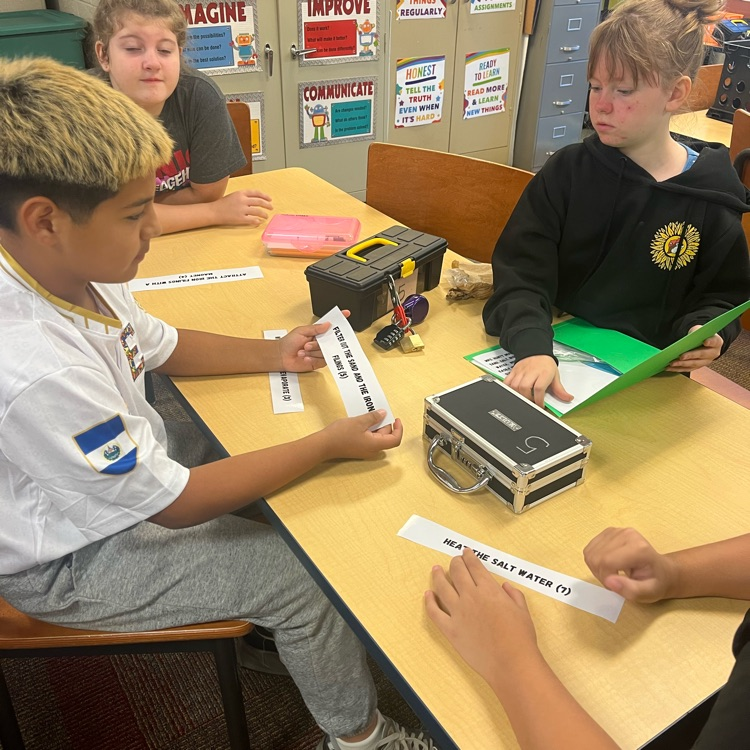 three students sit around a rectangular table and inspect science clues that will help them open locks on a lock box 