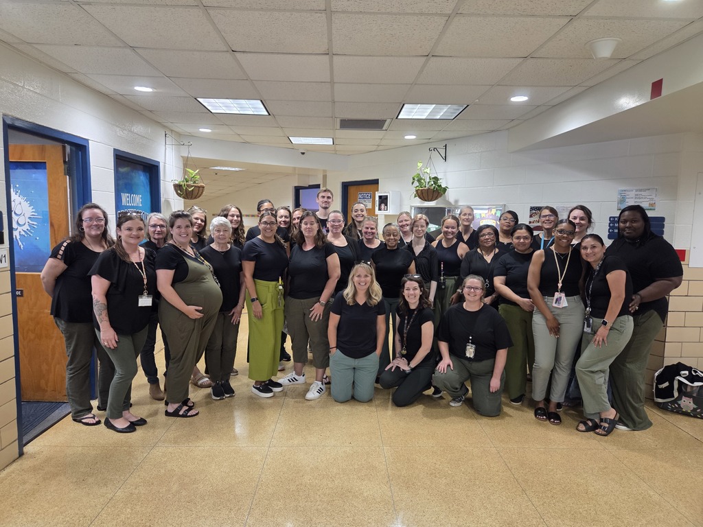 Image contains a staff posing in the front hallway.  Staff are all wearing black shirts and green pants for a twin spirit day.