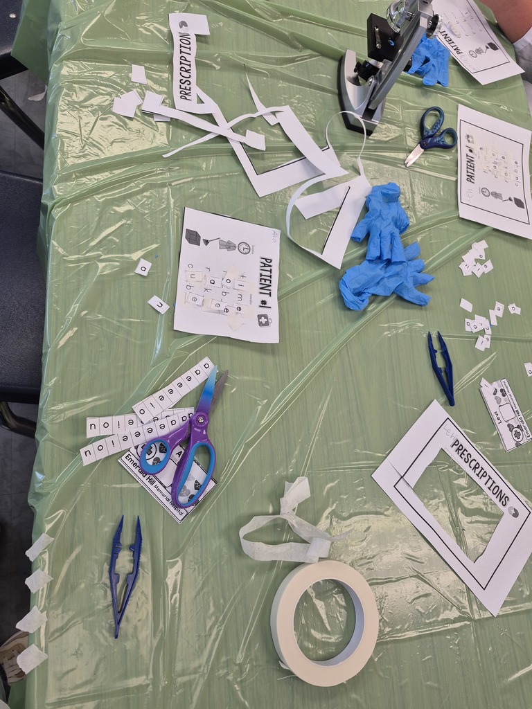 “Second grade students in Mrs. Bridges’ class participate in a Silent E Surgery literacy activity. Students wear white lab coats, safety goggles, and gloves while working with patient files, prescriptions, and word cards at tables set up like an operating room. Smiling children pose with their materials, and the classroom is decorated with lab coats, microscopes, and learning tools.