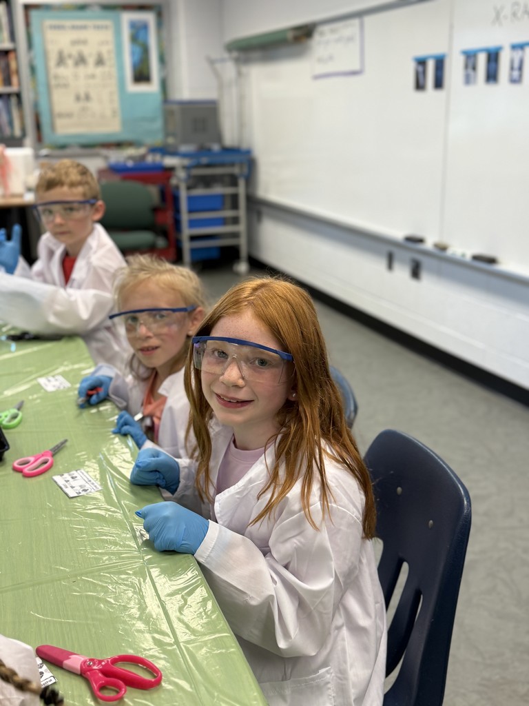 “Second grade students in Mrs. Bridges’ class participate in a Silent E Surgery literacy activity. Students wear white lab coats, safety goggles, and gloves while working with patient files, prescriptions, and word cards at tables set up like an operating room. Smiling children pose with their materials, and the classroom is decorated with lab coats, microscopes, and learning tools.