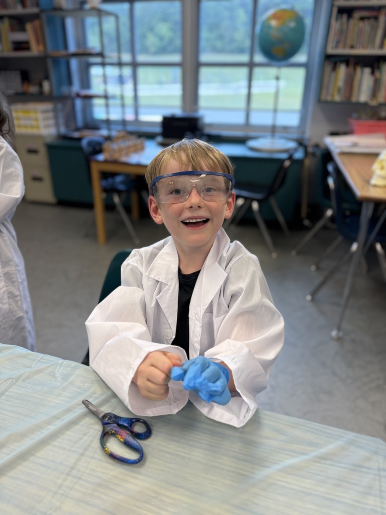 “Second grade students in Mrs. Bridges’ class participate in a Silent E Surgery literacy activity. Students wear white lab coats, safety goggles, and gloves while working with patient files, prescriptions, and word cards at tables set up like an operating room. Smiling children pose with their materials, and the classroom is decorated with lab coats, microscopes, and learning tools.