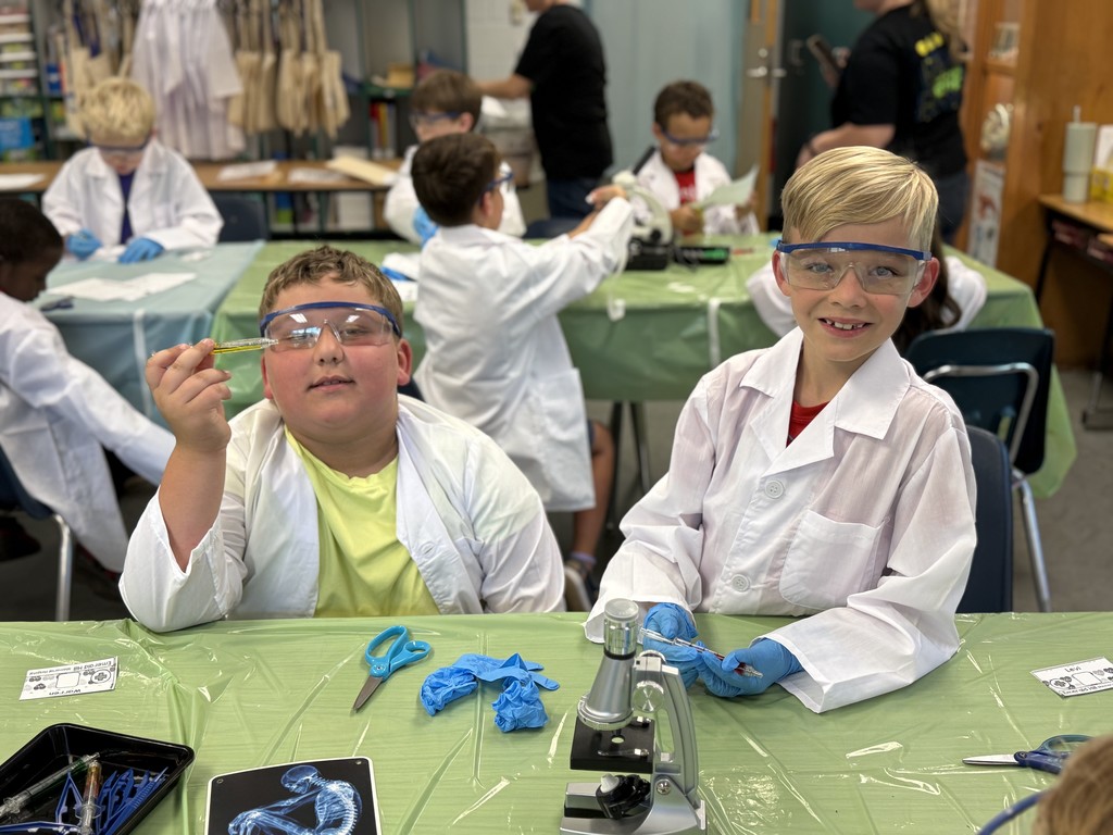 “Second grade students in Mrs. Bridges’ class participate in a Silent E Surgery literacy activity. Students wear white lab coats, safety goggles, and gloves while working with patient files, prescriptions, and word cards at tables set up like an operating room. Smiling children pose with their materials, and the classroom is decorated with lab coats, microscopes, and learning tools.