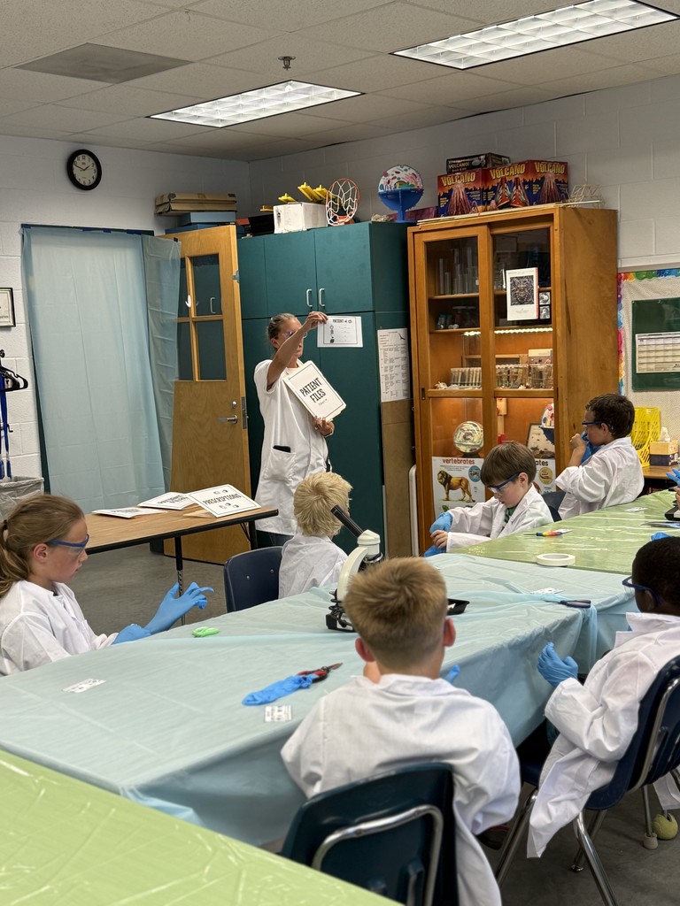 “Second grade students in Mrs. Bridges’ class participate in a Silent E Surgery literacy activity. Students wear white lab coats, safety goggles, and gloves while working with patient files, prescriptions, and word cards at tables set up like an operating room. Smiling children pose with their materials, and the classroom is decorated with lab coats, microscopes, and learning tools.