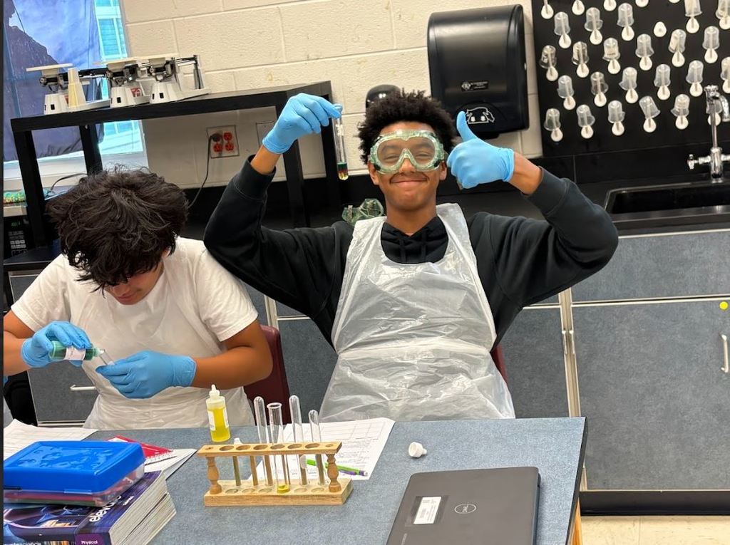 Two students work on a science experiment in a classroom. Both are wearing gloves and aprons; one student also has on safety goggles and holds up a test tube with a smile and thumbs up. The other student is focused on pouring liquid into a container. A test tube rack, laptop, and lab supplies are on the table.