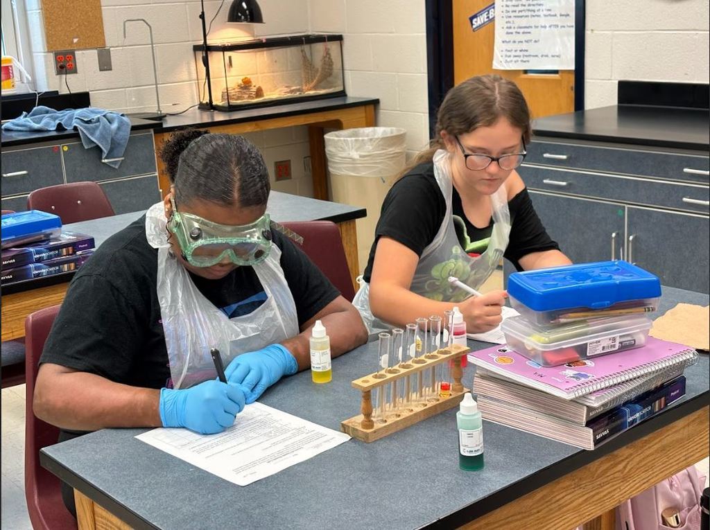 Two students in a science classroom work on a lab activity. Both are wearing aprons and gloves, with one student also wearing safety goggles. They are writing on worksheets at their desks, which have test tubes in a wooden rack, lab supplies, and notebooks. 