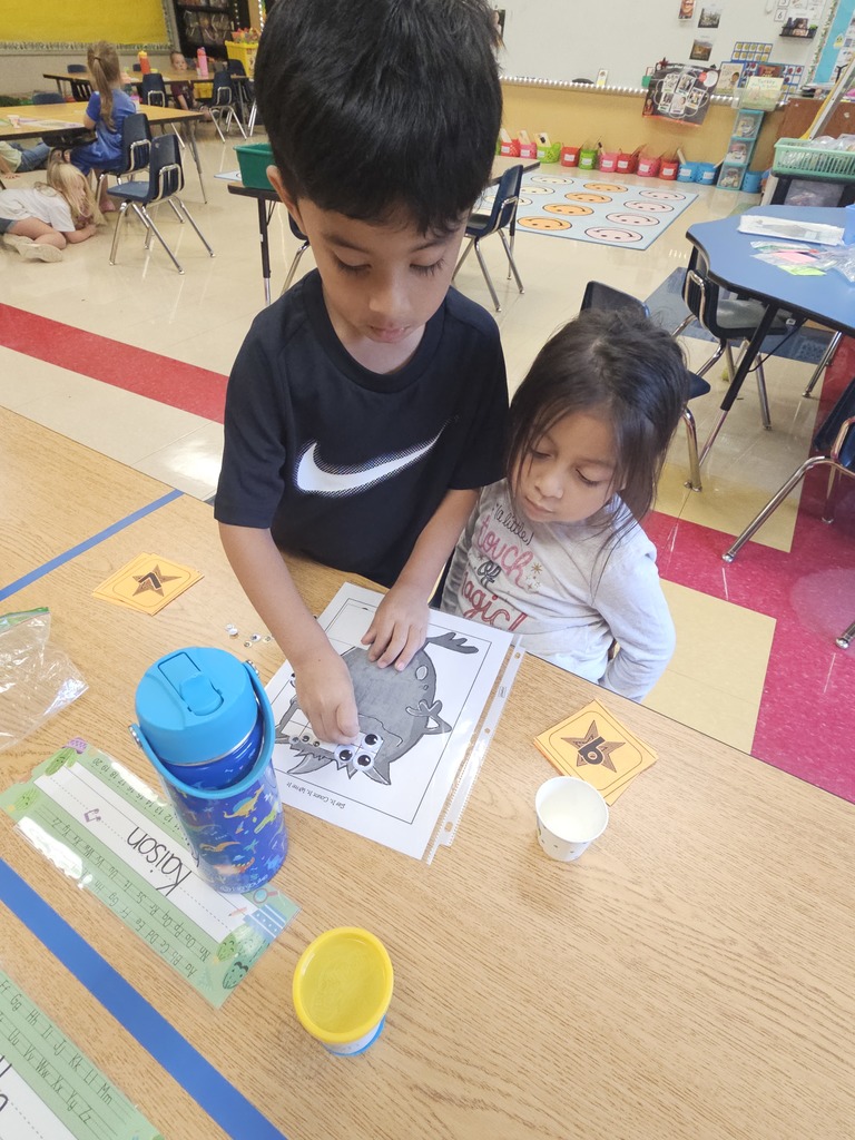 A boy and girl sit at a table together playing the monster eyeball game. The boy places googly eyes on the monster worksheet while the girl looks on. Their supplies, including a water bottle and Play-Doh container, are on the desk.