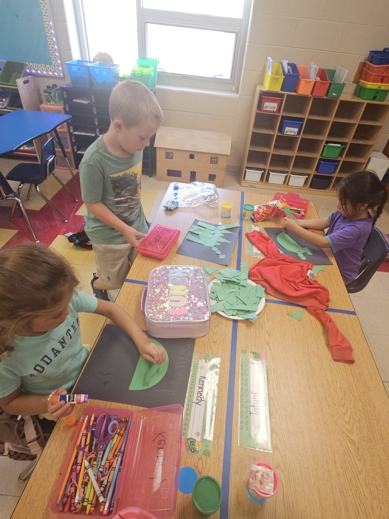 Three kindergarten students sit at a table with paper, crayons, and school supplies. They are focused on gluing and arranging green paper cutouts on black paper to create shape dinosaurs.