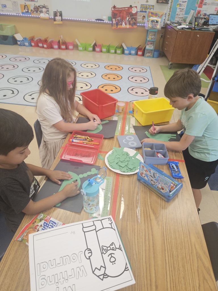 Three students sit at a table with supplies, crayons, and a writing journal on the desk. They are tracing and gluing paper shapes to make dinosaurs. A colorful rug with smiley faces is visible in the background.