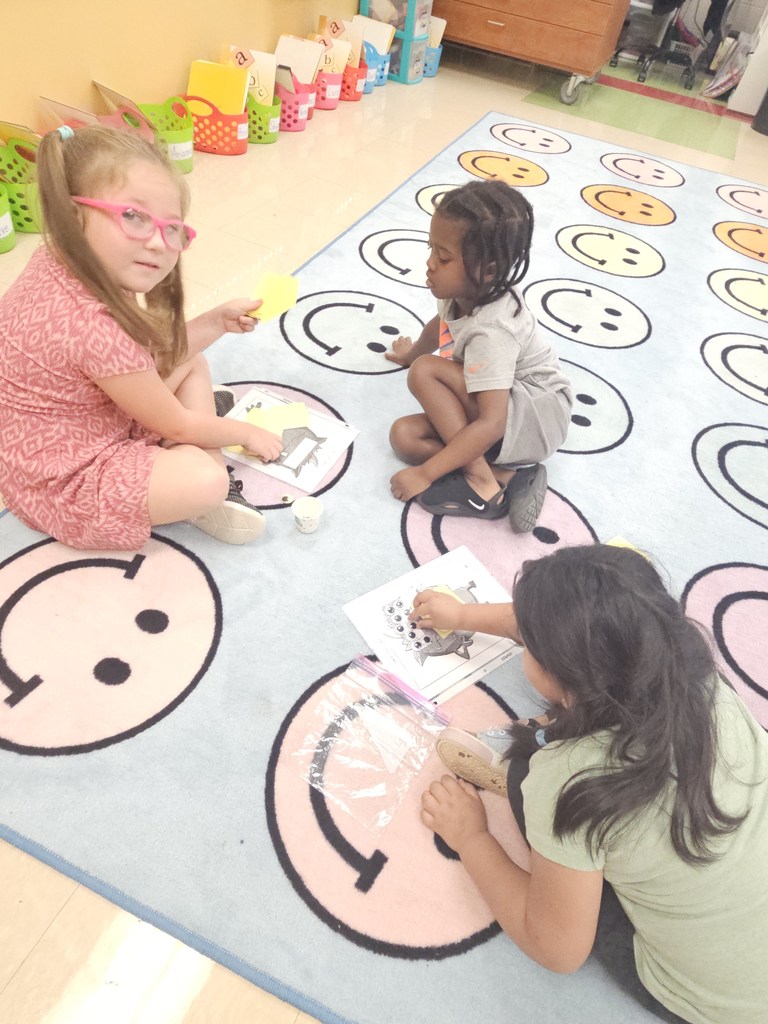 Three students sit together on a rug decorated with smiley faces, playing the monster eyeball game. One girl holds a number card while another places googly eyes onto the monster worksheet.