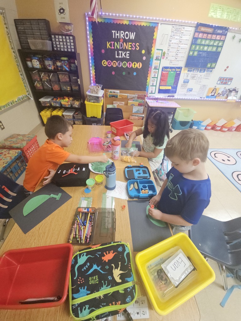 Three kindergarten students sit at a classroom table covered with supplies, lunchboxes, and crayons. They are gluing large green paper shapes onto black construction paper as part of a shape-building activity. A bulletin board behind them reads “Throw Kindness Like Confetti.”