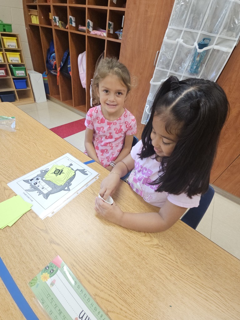 Two girls sit at a table playing the monster eyeball counting game. One girl smiles at the camera while the other pulls a number card and prepares to place eyeballs on the monster sheet.