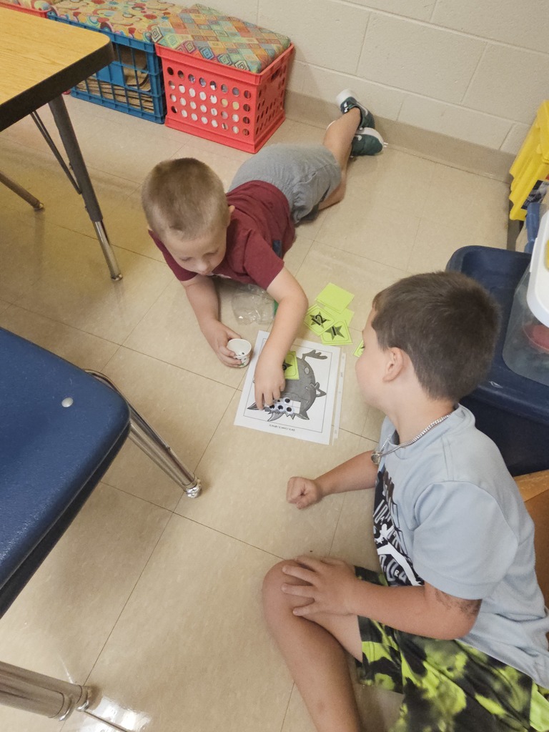 Two boys lie on the classroom floor, playing the “monster eyeball” counting game. One child holds a small cup with counters while the other places pieces on a worksheet with a cartoon monster.