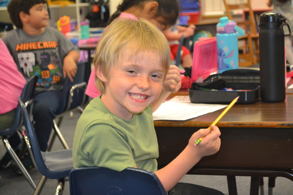 Image contains a smiling kindergartner at his desk in his classroom.  Student is holding a pencil.