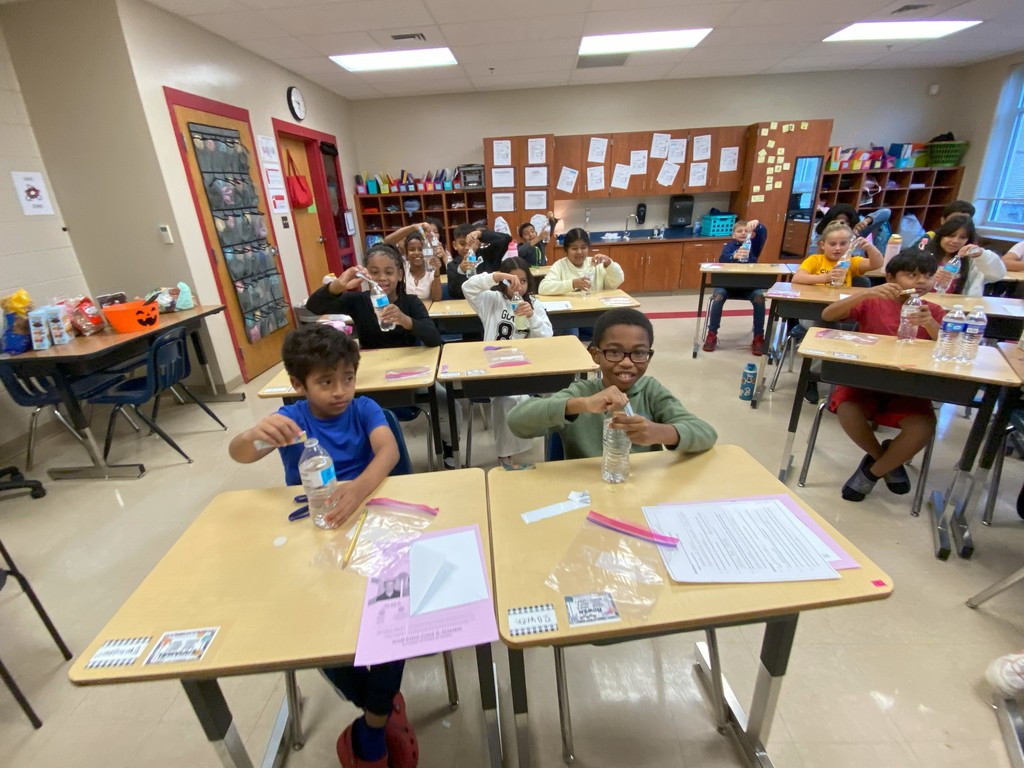 A classroom of students sitting at desks, each holding a water bottle and adding materials inside as part of a science or craft activity. The students look engaged and focused.