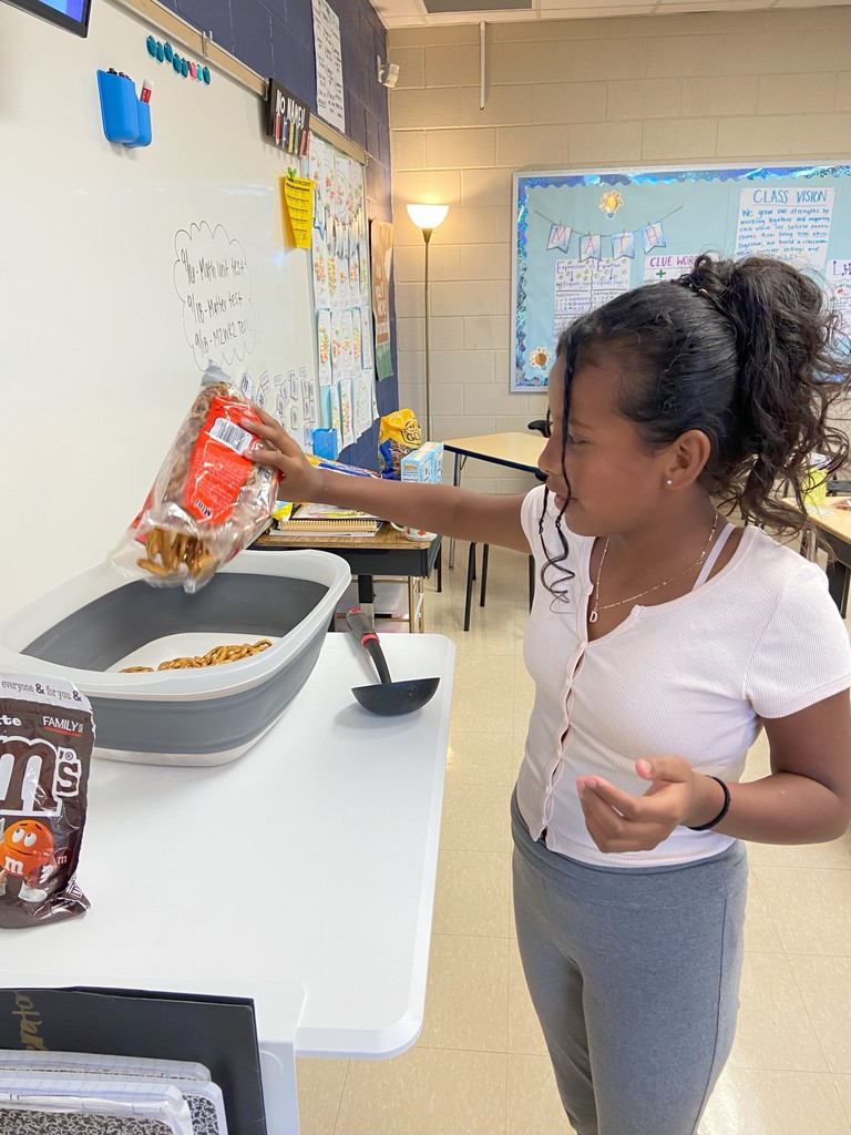 A student pours a bag of pretzels into a large gray container on a table, preparing a snack mix.