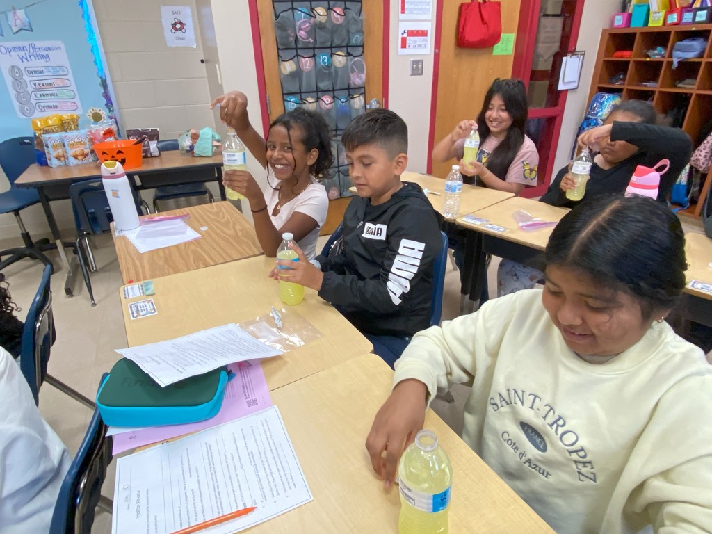 Several students smiling and showing water bottles filled with a yellow liquid mixture, demonstrating progress in their experiment.