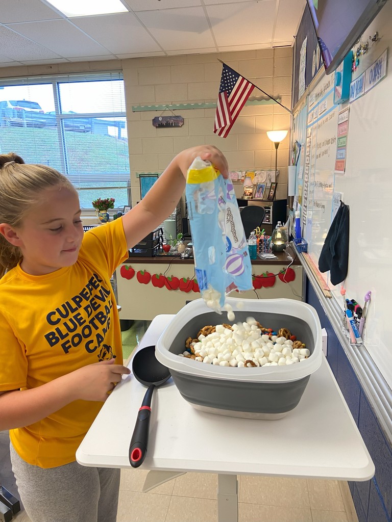 A student in a yellow shirt adds mini marshmallows from a bag into the same large container, which is already filled with pretzels and candy, as part of the snack mix preparation.
