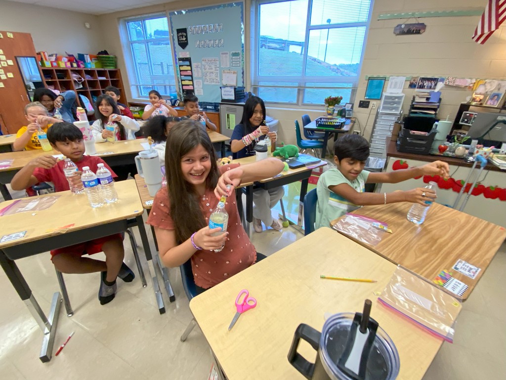 Smiling students at their desks, dropping items into water bottles while working on an activity. A girl in the front center holds up her bottle proudly while classmates around her participate.