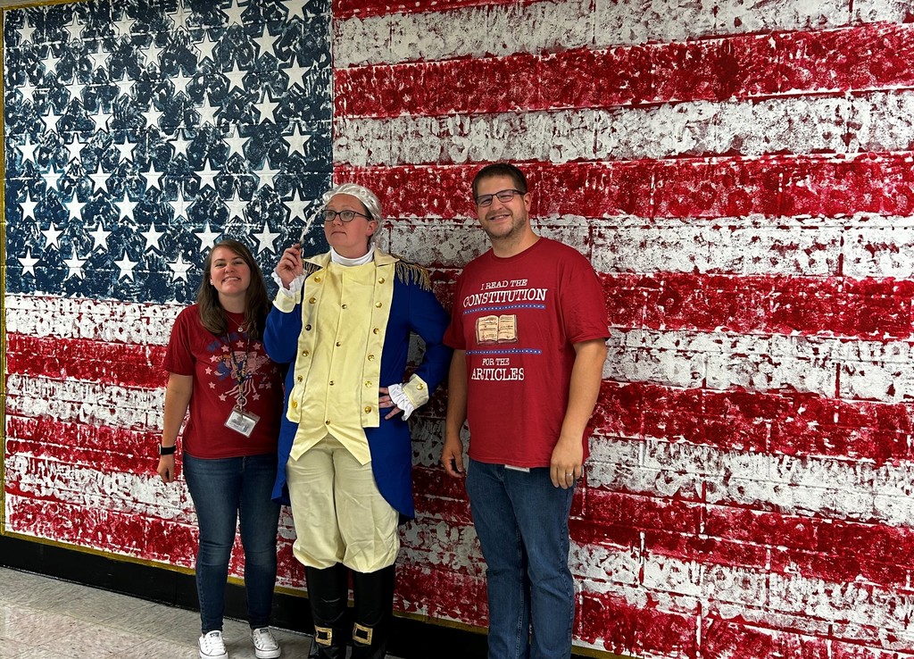 Three adults stand in front of a large painted American flag mural. One person in the center is dressed in a blue and yellow colonial-style costume with a white wig, while the two others wear red shirts with patriotic designs and text. All three are smiling and posing together.
