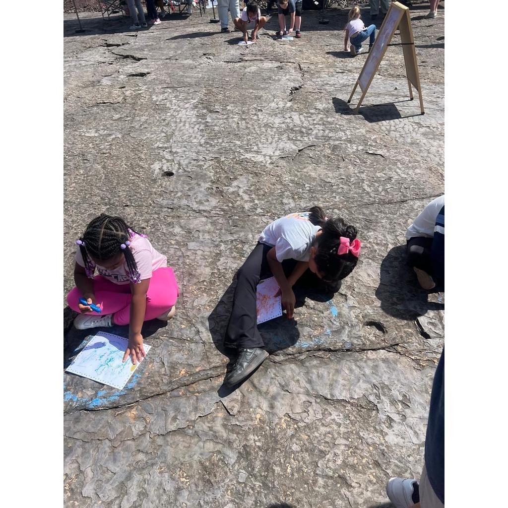 Students sit on rocky ground outside, coloring paper with crayons during a field trip.