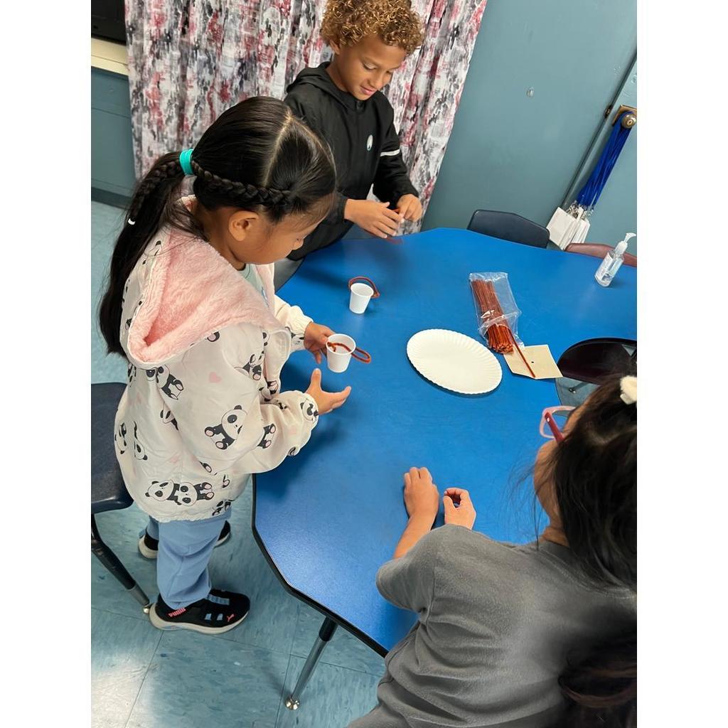 Three students gather at a blue table, building small parachute projects with cups and strings.