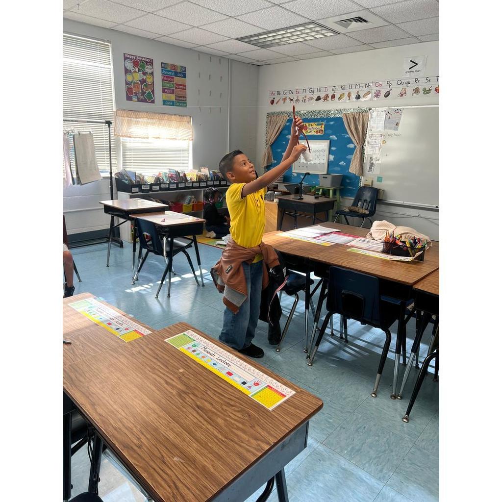 A student in a yellow shirt tests a homemade parachute by holding it high in a classroom.
