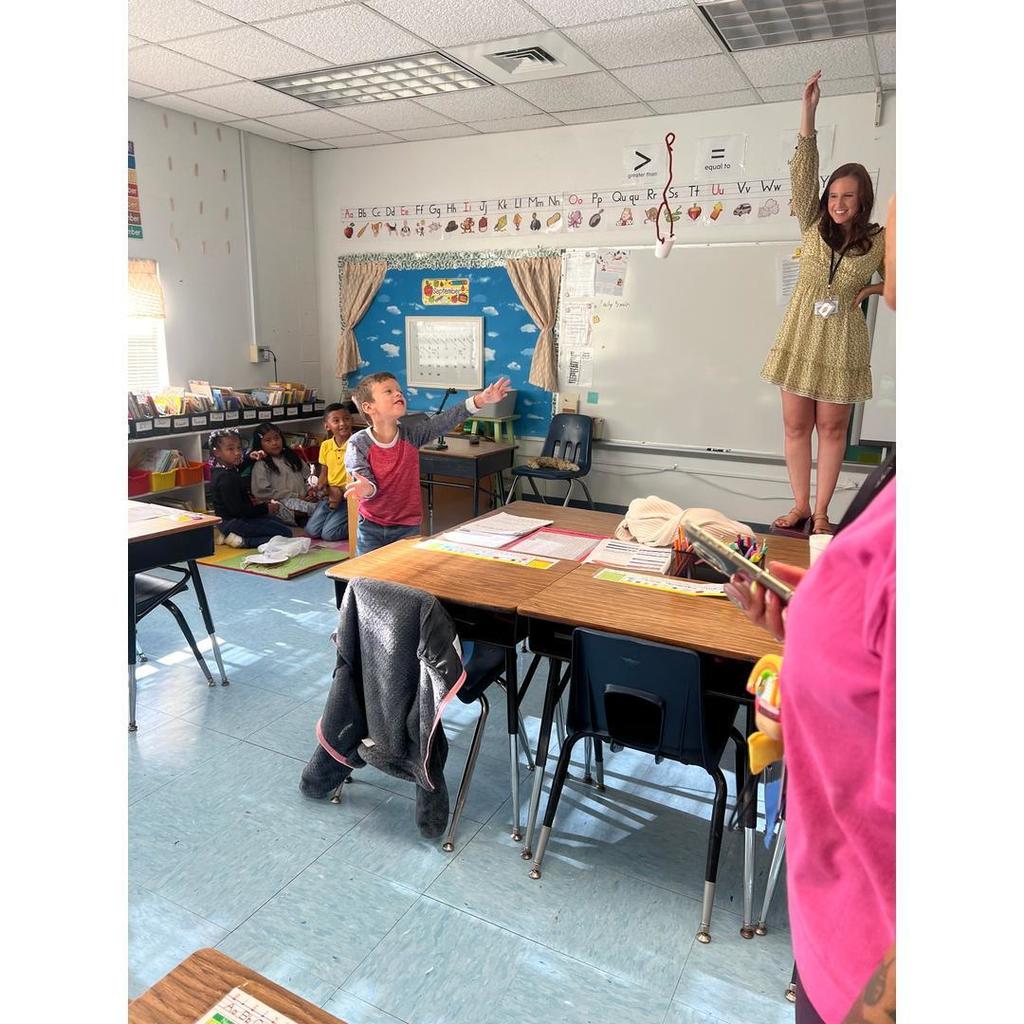 A teacher helps a student test a parachute project while classmates watch from the carpet.