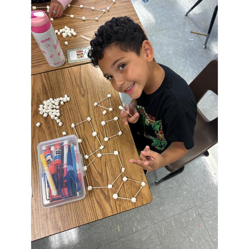 A student displays an elaborate marshmallow and toothpick design across the desk with a big smile.