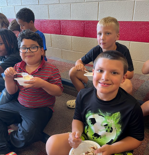 Several boys sit together against a wall, smiling and eating bowls of ice cream during a classroom treat.
