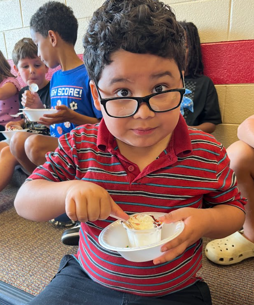 A boy in a red striped shirt and glasses looks up while scooping ice cream from a bowl with a spoon.