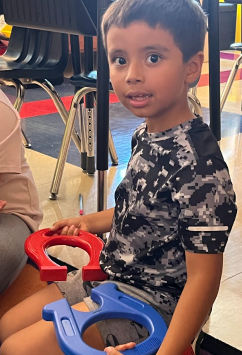 A boy in a camouflage shirt sits on the floor holding two large horseshoe magnets, one red and one blue, during a science activity.