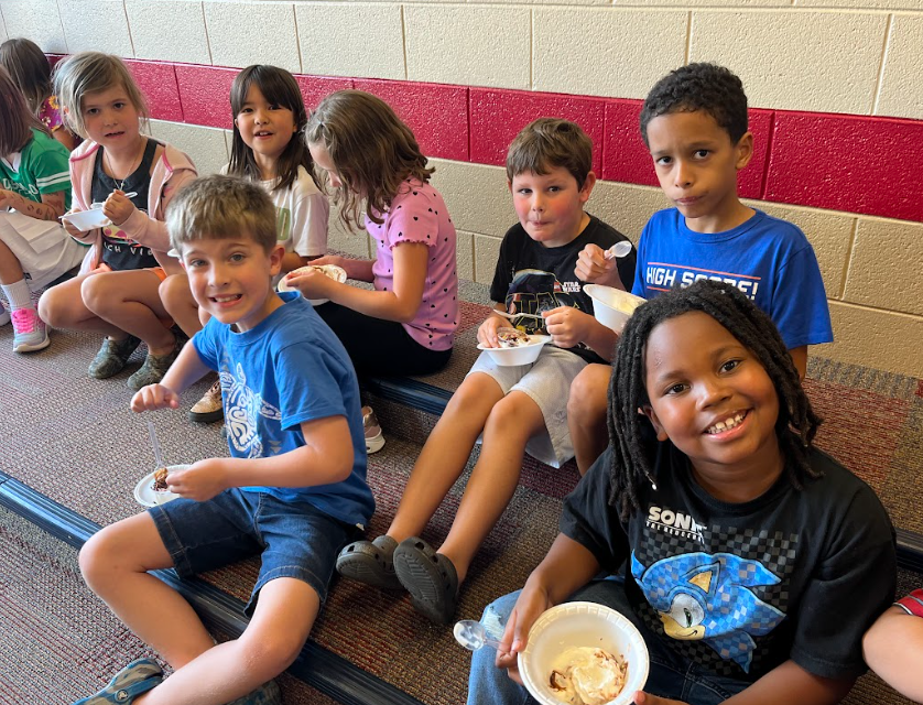 More students sit on risers with bowls of ice cream, some smiling at the camera while others eat their treat.