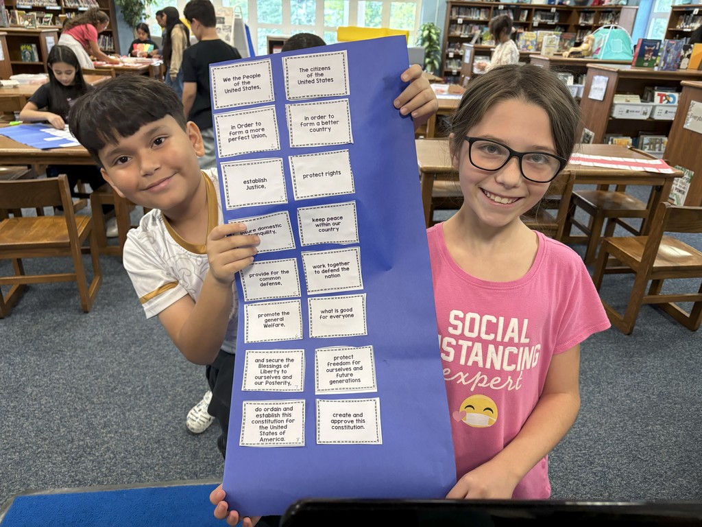 Image contains one boy, one girl holding a blue piece of paper with glued pieces about the Preamble in the school library.