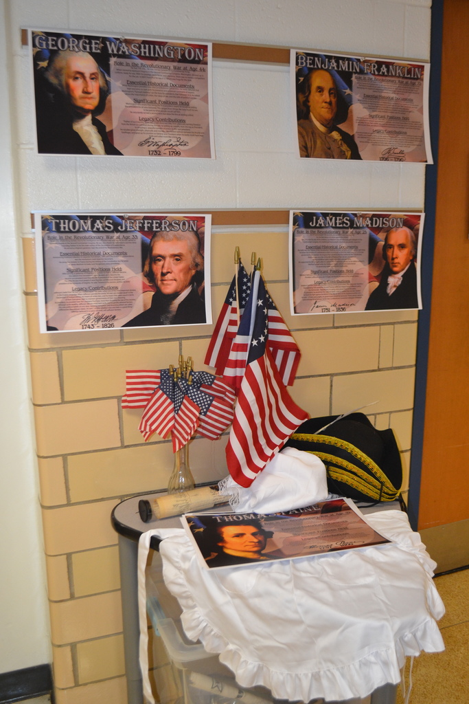 Image contains a display of donated educational materials - sign on George Washington James Madison Thomas Jefferson Benjamin Franklin.  There are a variety of American flags and colonial era style clothing pieces.  This is located in a school hallway.