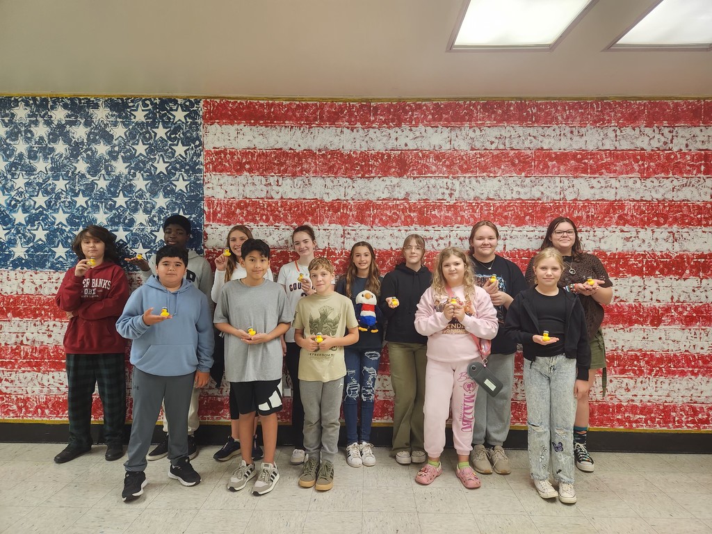 A group of middle school students stand in front of a large painted American flag mural. Many are holding small patriotic rubber ducks, while one student in the center holds a plush eagle. The students are smiling and posing together to celebrate Constitution Day.