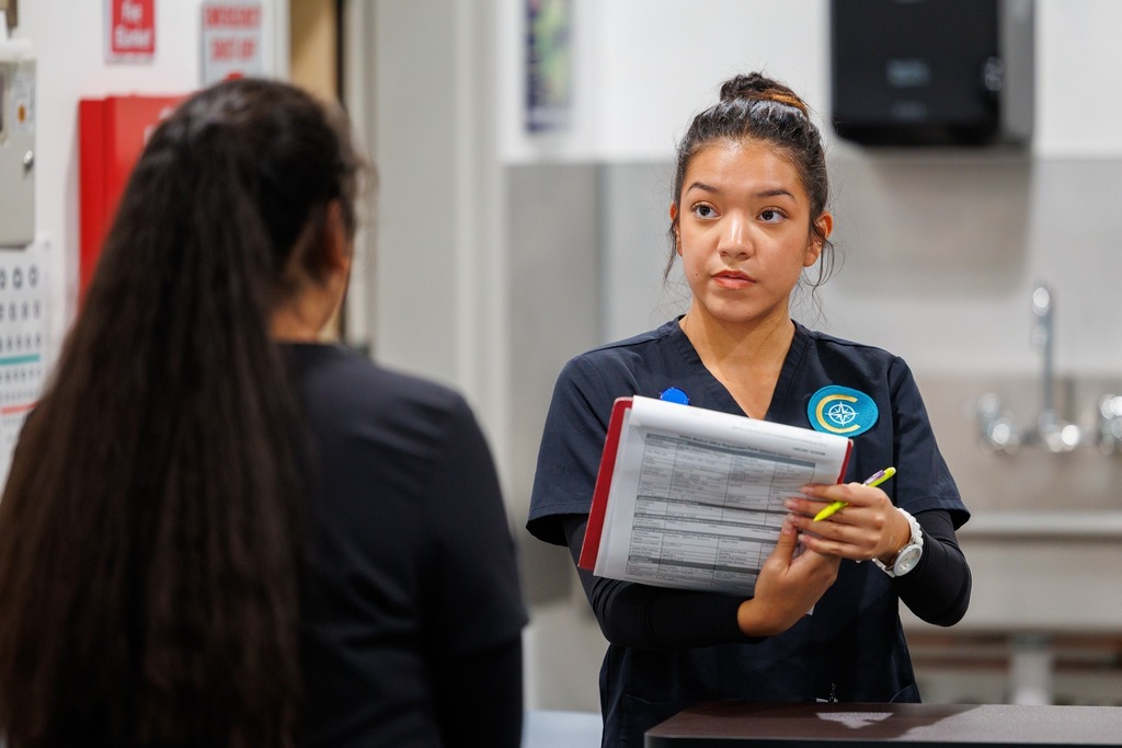 A CTEC medical assistant student holds a clipboard and yellow pen, explaining notes to a classmate while practicing rooming procedures in a medical lab.