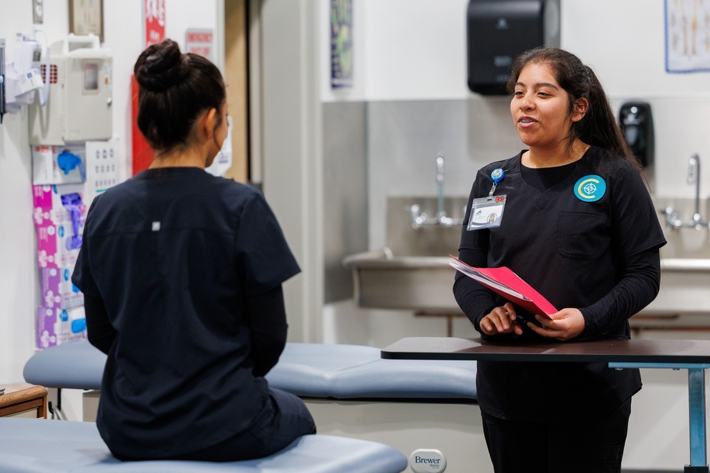 A student stands with a clipboard, smiling while speaking to another student seated on an exam table during a practice patient intake scenario.