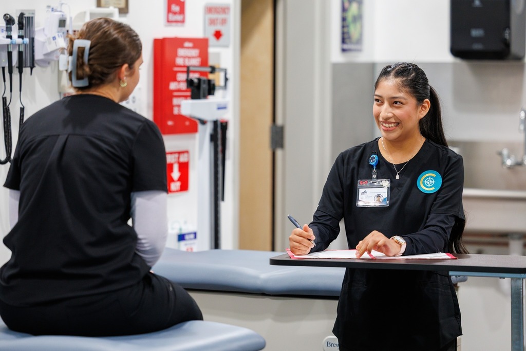 A student stands with a clipboard, writing notes while smiling at a classmate seated on an exam table during rooming skills practice.