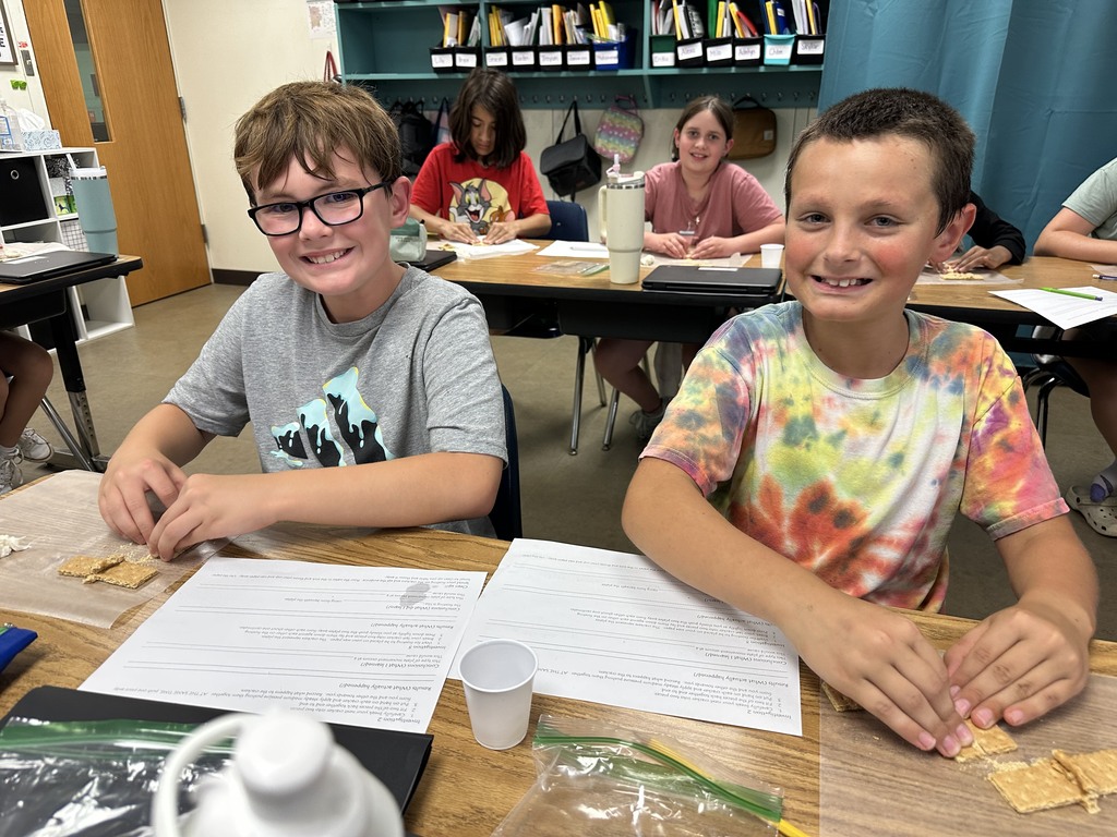 Two fifth-grade students sit at their desks smiling at the camera while working with graham crackers on wax paper. Other students can be seen in the background also participating in the activity.