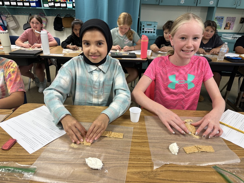 Two fifth-grade students sit side by side at their desks, smiling at the camera while using graham crackers and frosting for a plate tectonics activity. Classmates in the background are engaged in the same project.