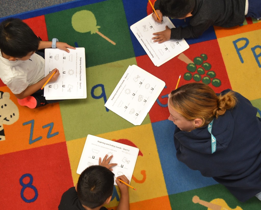 Arial photo of 3 students working on a brightly colored carpet with their teacher.  Each as a paper learning about the letter Uu.
