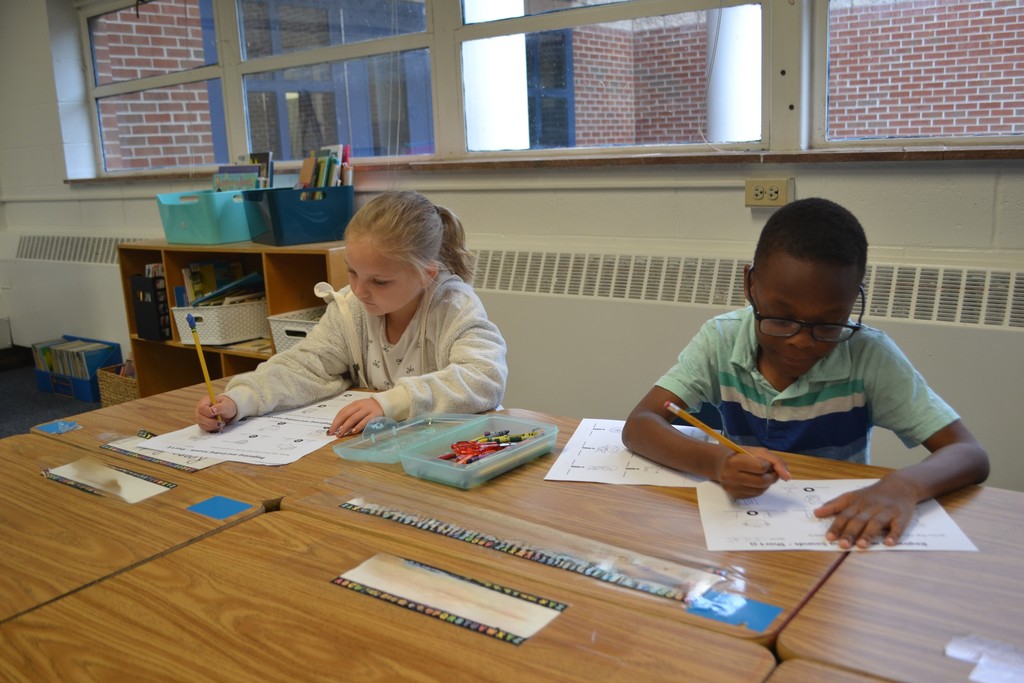 Image contains a girl and boy sitting at their desks working on their reading skill.