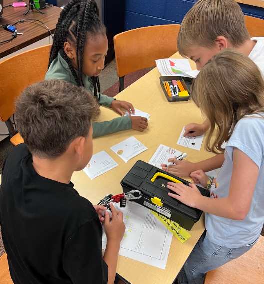 Four students collaborate at a table with papers, cards, and a locked black toolbox. They lean in closely, reading and discussing clues.