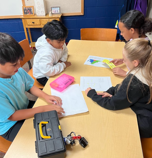 Four students sit at a table studying worksheets and a map. A locked black toolbox with a red lock is in front of them. They appear to be solving a puzzle together.