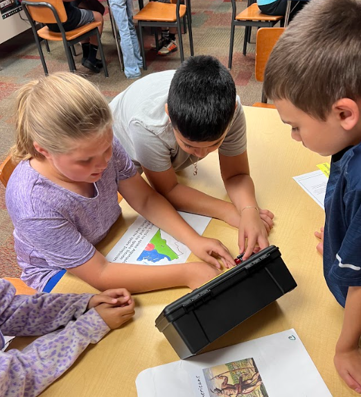 Three students gather around a table, working together to open a locked black box. A map worksheet and other papers are spread out on the table.